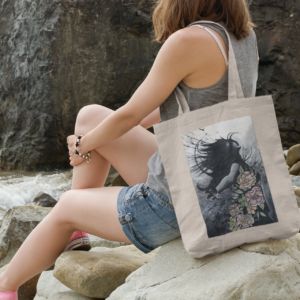 Woman sitting on rocky shoreline with a natural canvas tote bag featuring an artistic illustration.