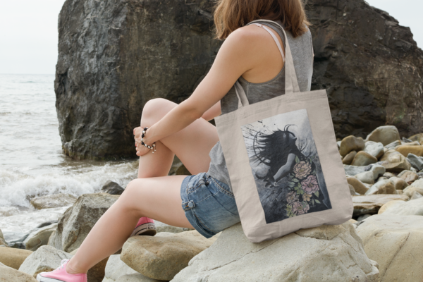 Woman sitting on rocky shoreline with a natural canvas tote bag featuring an artistic illustration.