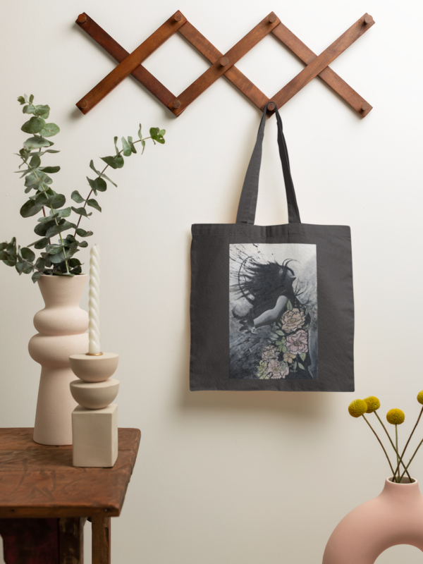 Black cotton tote bag with artistic design, displayed on a wooden wall hook.